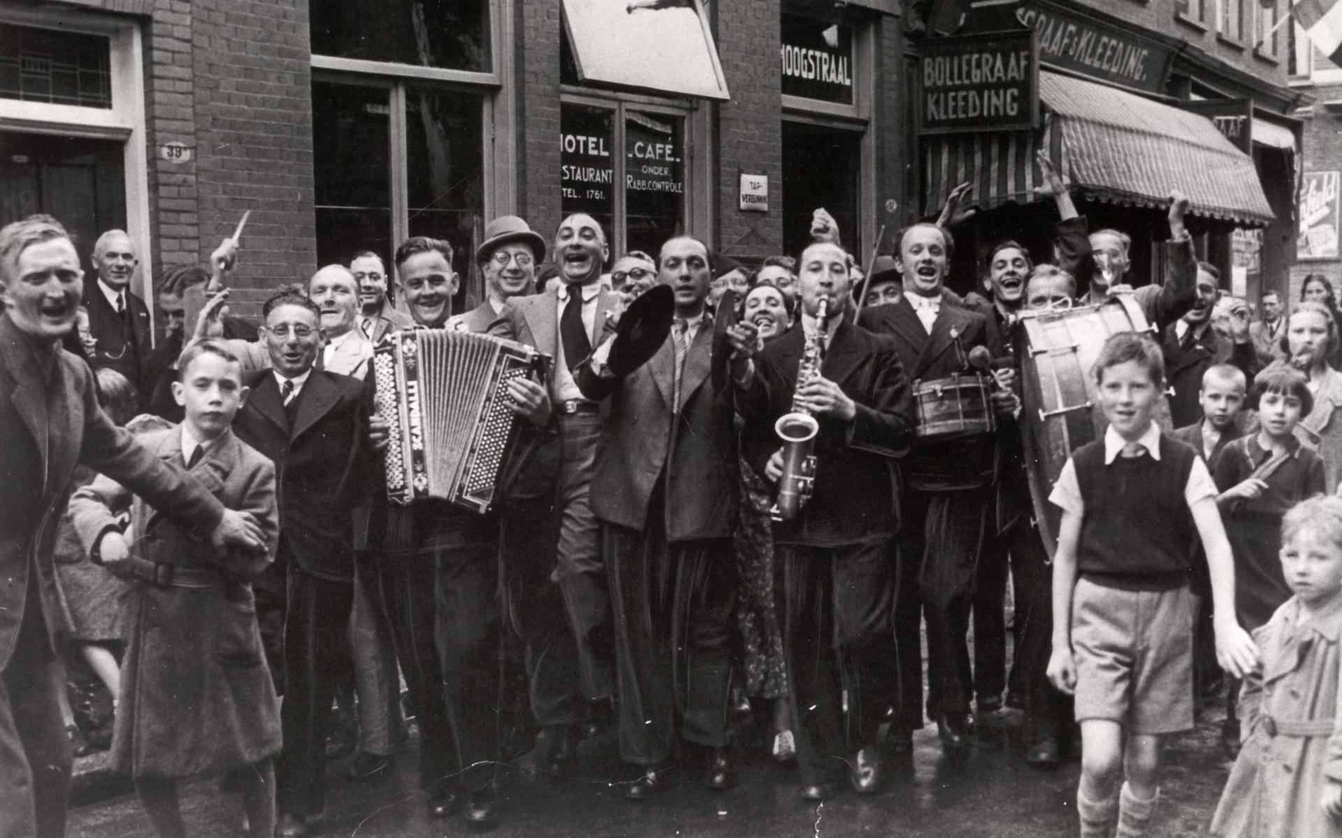 The Bolly Band tijdens een optreden in de Folkingestraat. Levie Bollegraaf (met stropdas) lacht onhoorbaar een bulderende lach, Jacob, de spil van de band, houdt de bekkens vast en broer Ruben blaast op de saxofoon . Op de achtergrond de kledingzaak van hun vader Simon Bollegraaf op Folkingestraat 35. 